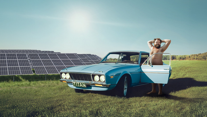 Ein Mann steht barfuß neben einer offenen blauen Autotür und entspannt sich neben Solarmodulen auf einer Wiese. - © PMT 2025