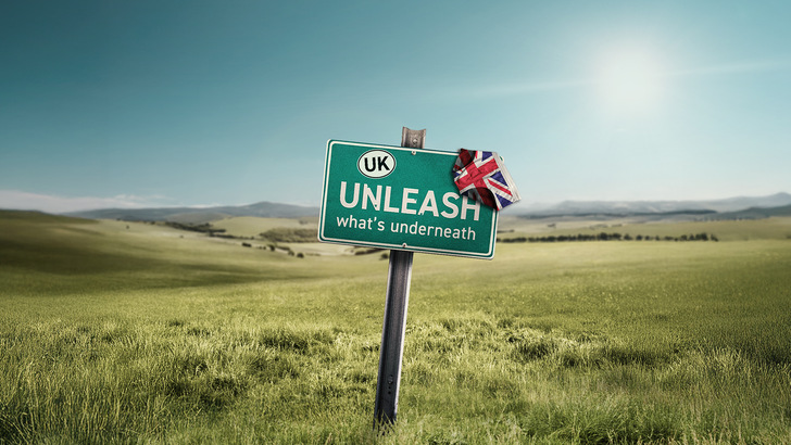 Auf einem Straßenschild auf einer grünen Wiese steht „UK Unleash what's beneath“ (Großbritannien, entfesseln Sie, was darunter ist), und über der Ecke hängt ein Union Jack. - © PMT 2025