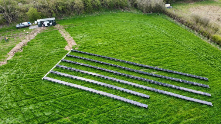 The solar plant stands on a meadow behind a row of trees. Next to it is the container with the storage unit and the power electronics.