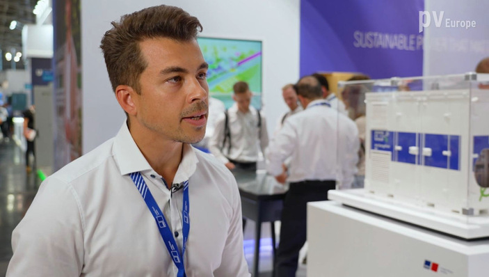 Man speaking at an exhibition booth with a model display, wearing a white shirt and lanyard. - © Vorsatz Media