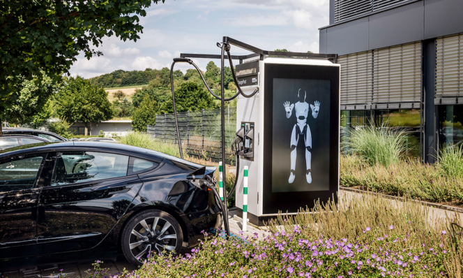 Electric car charging at a station with a robot image, near a building and greenery under a clear sky. - © ADS Tec Energy