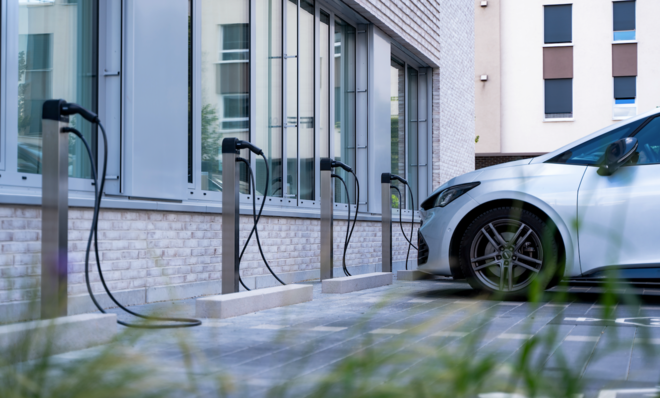 White electric car charging at a station outside a building, with multiple charging units visible. - © Mahle ChargeBig