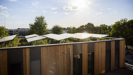 Solar panels on a rooftop with wooden siding, surrounded by trees and bathed in sunlight. - © Bauer Solar
